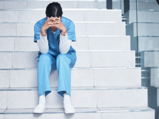 A woman in blue scrubs sits on hospital steps with her head in her hands.